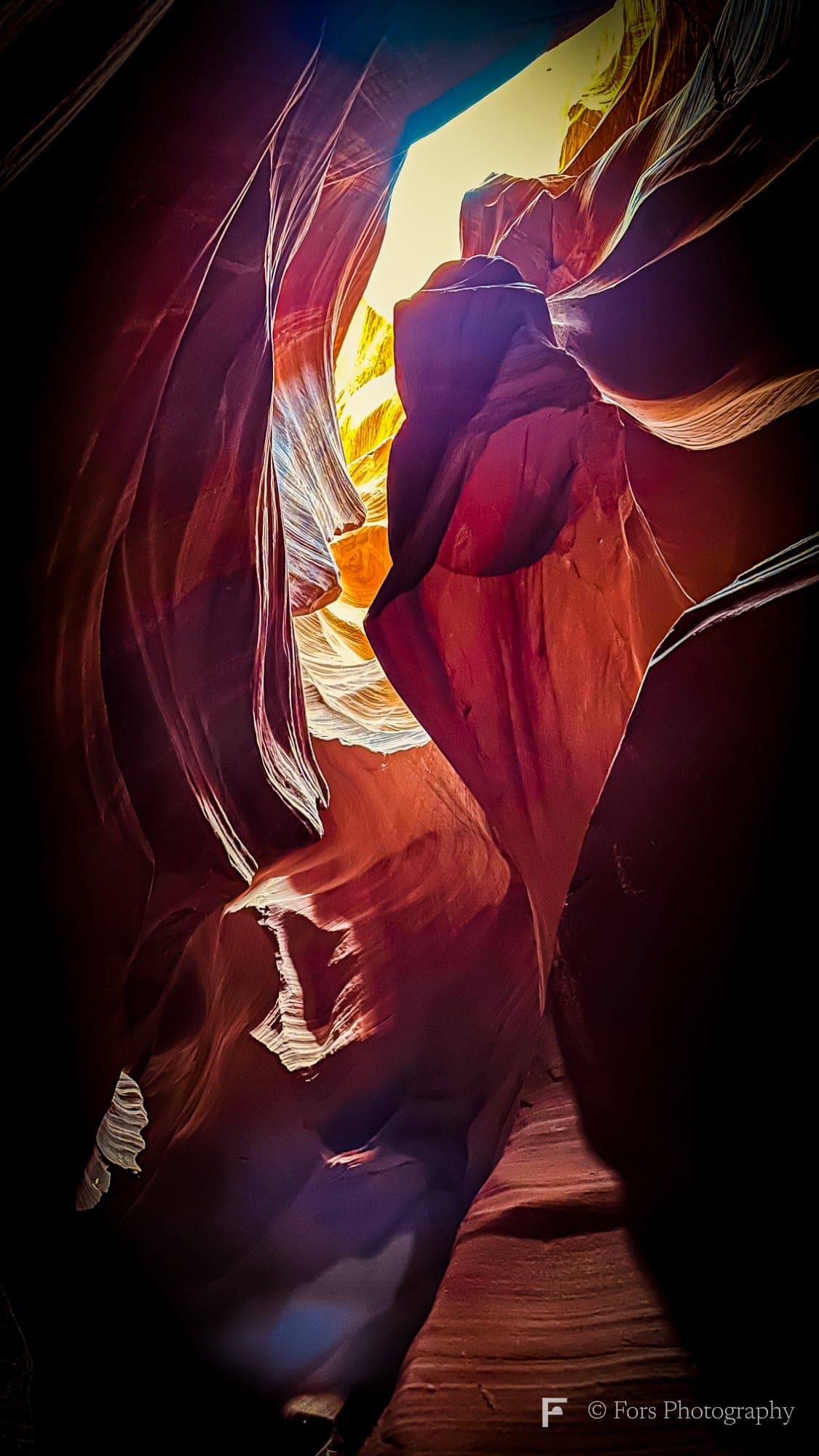 A narrow, winding slot canyon seen from below, with deep red and purple sandstone walls in shadow around a bright yellow opening of sky at the top.