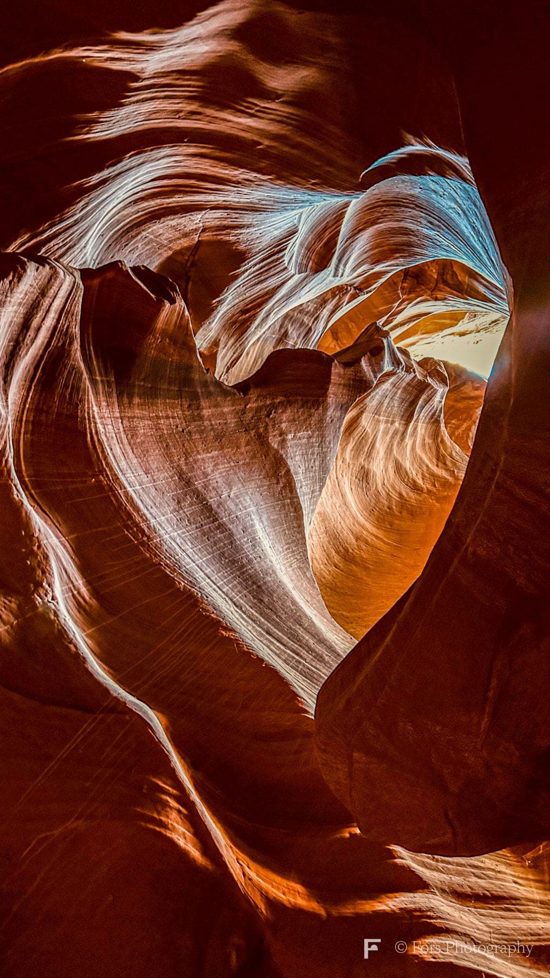 Looking up inside a sandstone slot canyon where the curving walls form a heart shape, lit in deep oranges and reds with a sliver of sky at the top.