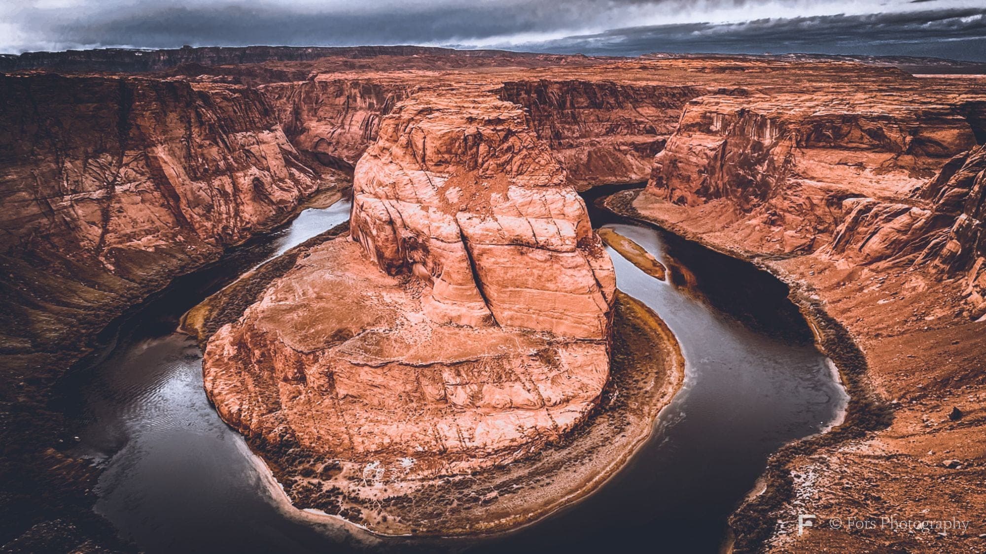 Aerial-style view of a river making a tight horseshoe-shaped bend around a tall sandstone mesa, under a dark bank of cloud.