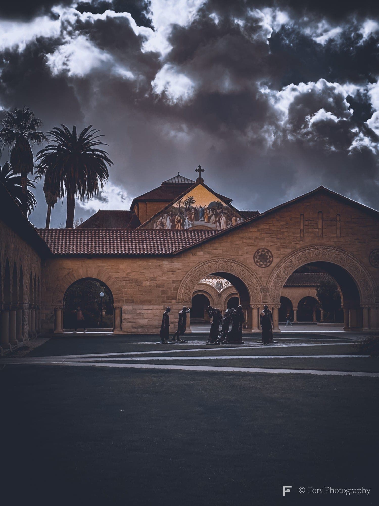 A sandstone arcade and a church front with a golden mosaic gable at Stanford, a group of dark sculpted figures in the courtyard, palm trees on the left and heavy storm clouds above.