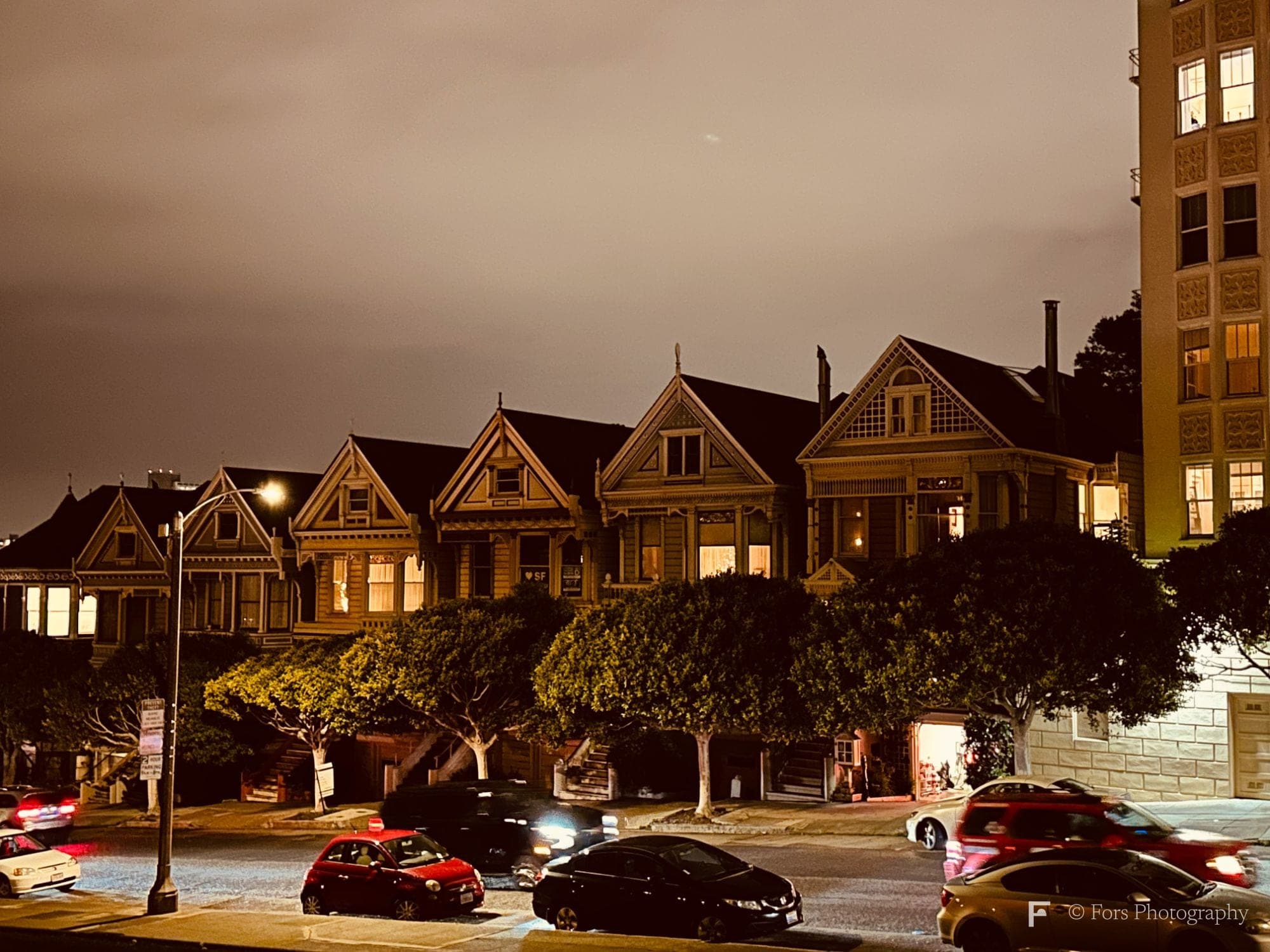 A row of Victorian houses at night with warmly lit windows and a trimmed line of trees in front; cars blur past on the street below under a sodium-orange sky.