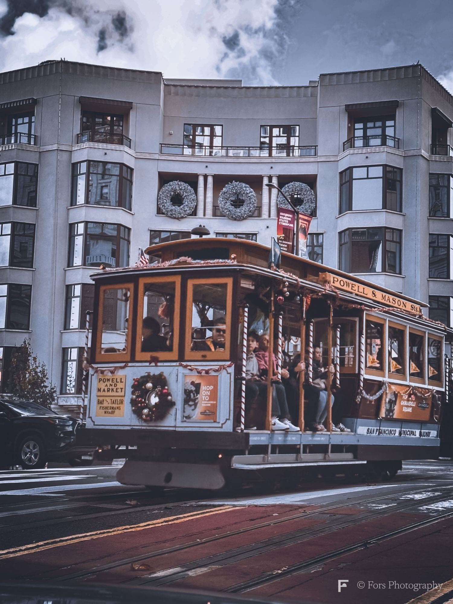 A San Francisco cable car decorated with Christmas wreaths and garlands, passengers standing on its running boards, in front of a pale curved building.