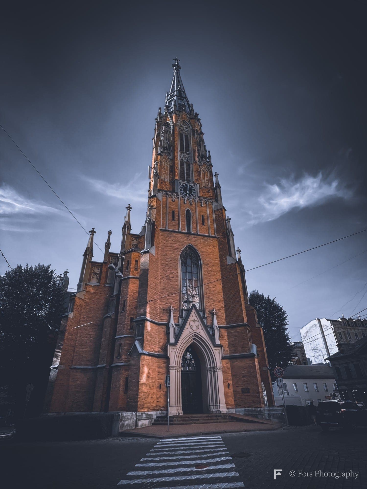 A tall red-brick Gothic church with a pointed spire, photographed from a pedestrian crossing in front, the brick glowing warm under a deep blue evening sky.
